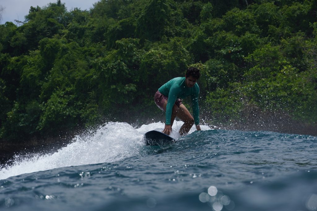 A man wearing a green Surf Journey Bali shirt surfing on gentle Balangan waves, set against a backdrop of a lush green cliff.