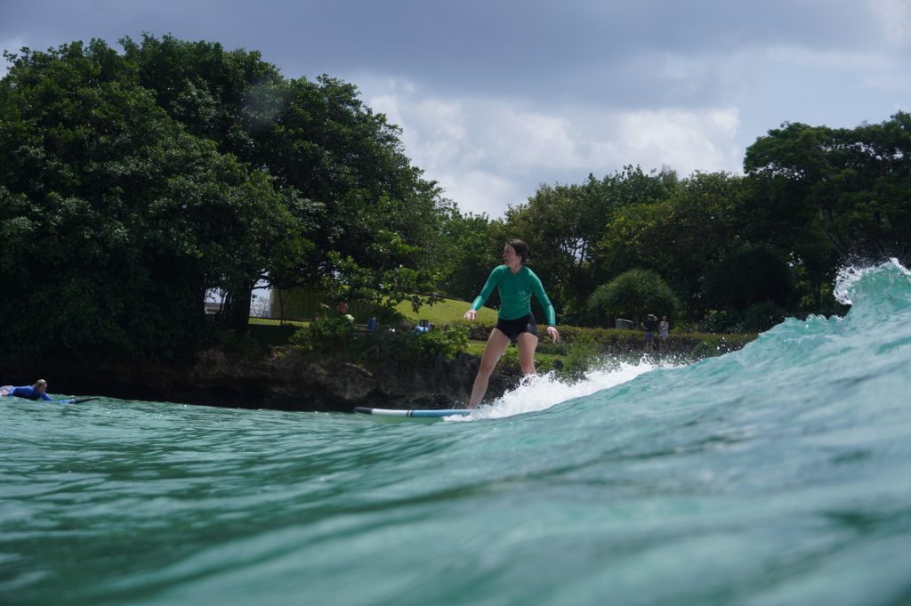 Blackstone Beach (Nusa Dua) - Surf Journey Bali Woman standing on the surfboard riding the gentle wave at Blackstone Beach (Nusa Dua) with Surf Journey Bali